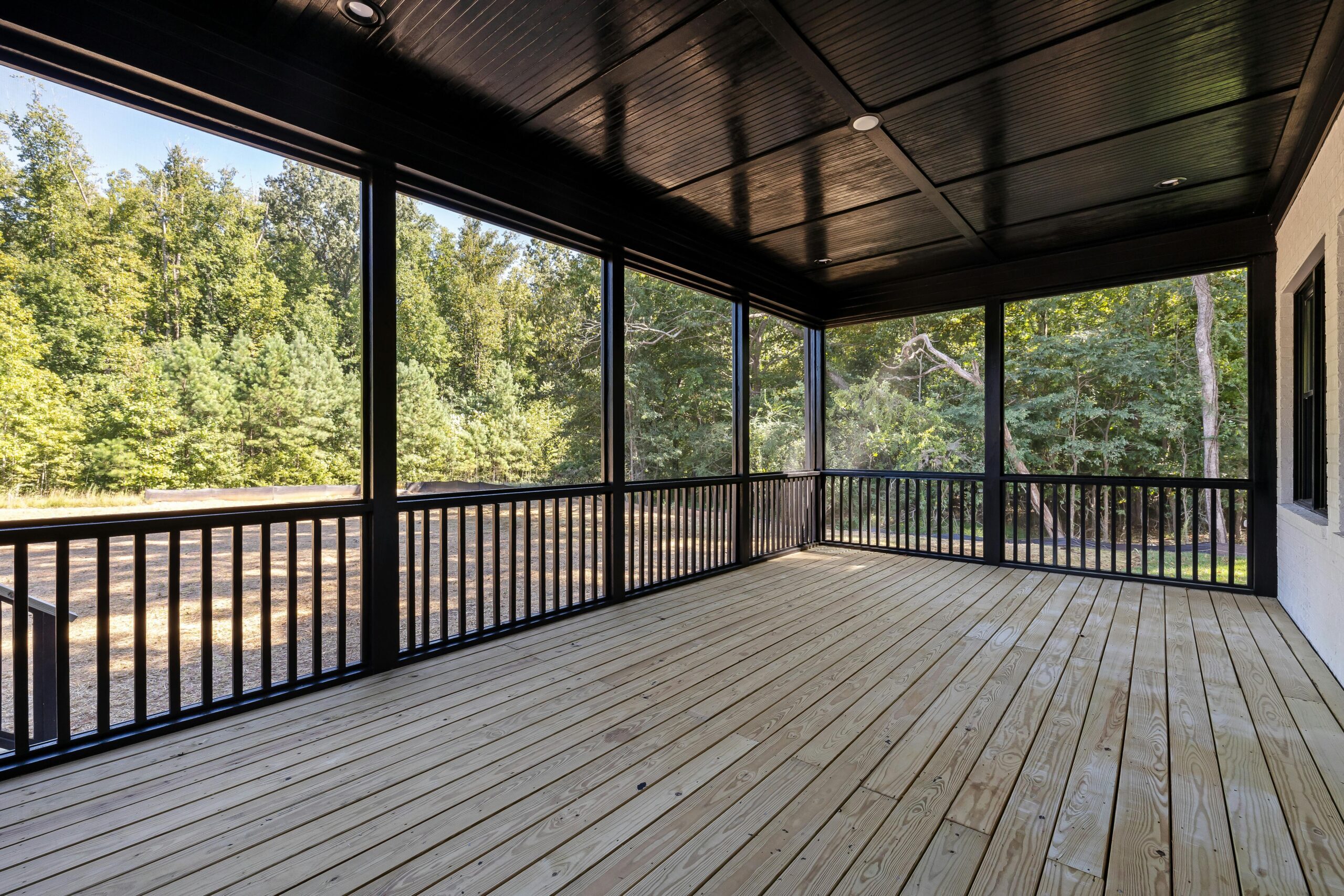 A spacious covered wooden deck with black railings and screened enclosures overlooking a lush, green forest.