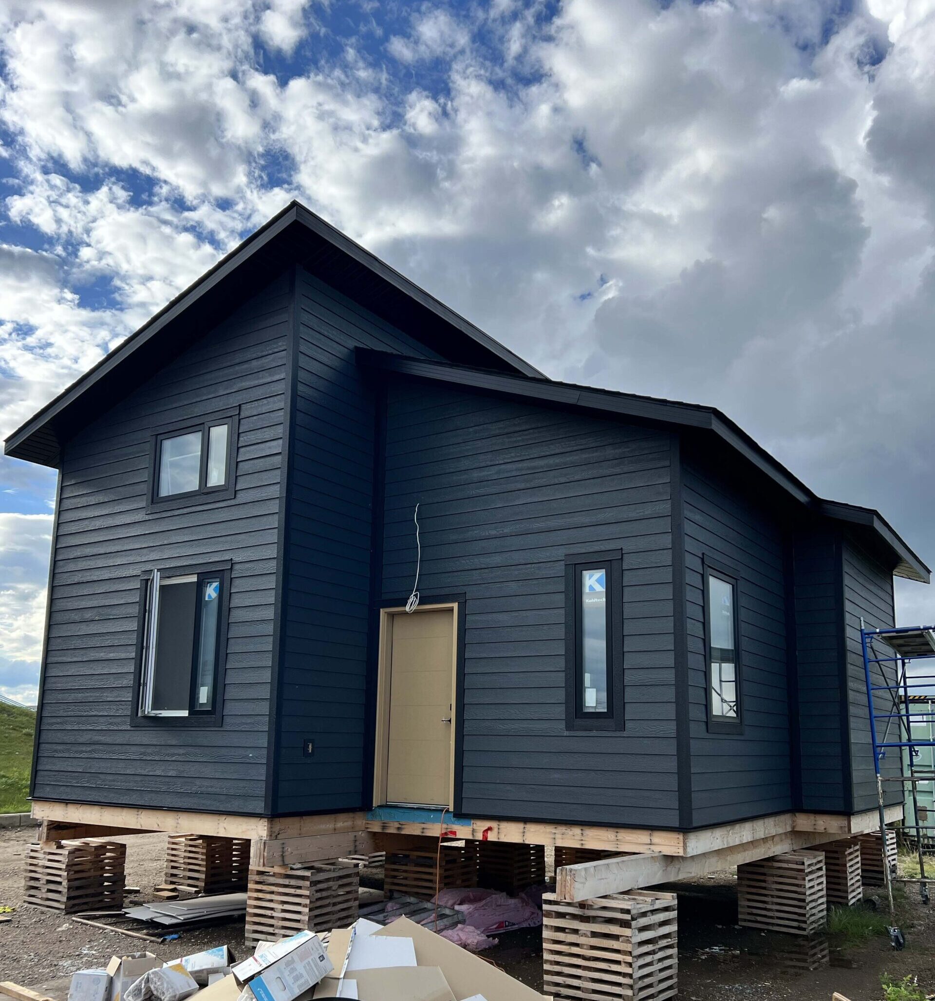 A modern house under construction with dark blue vinyl siding, large windows, and a beige door, elevated on temporary wooden blocks.