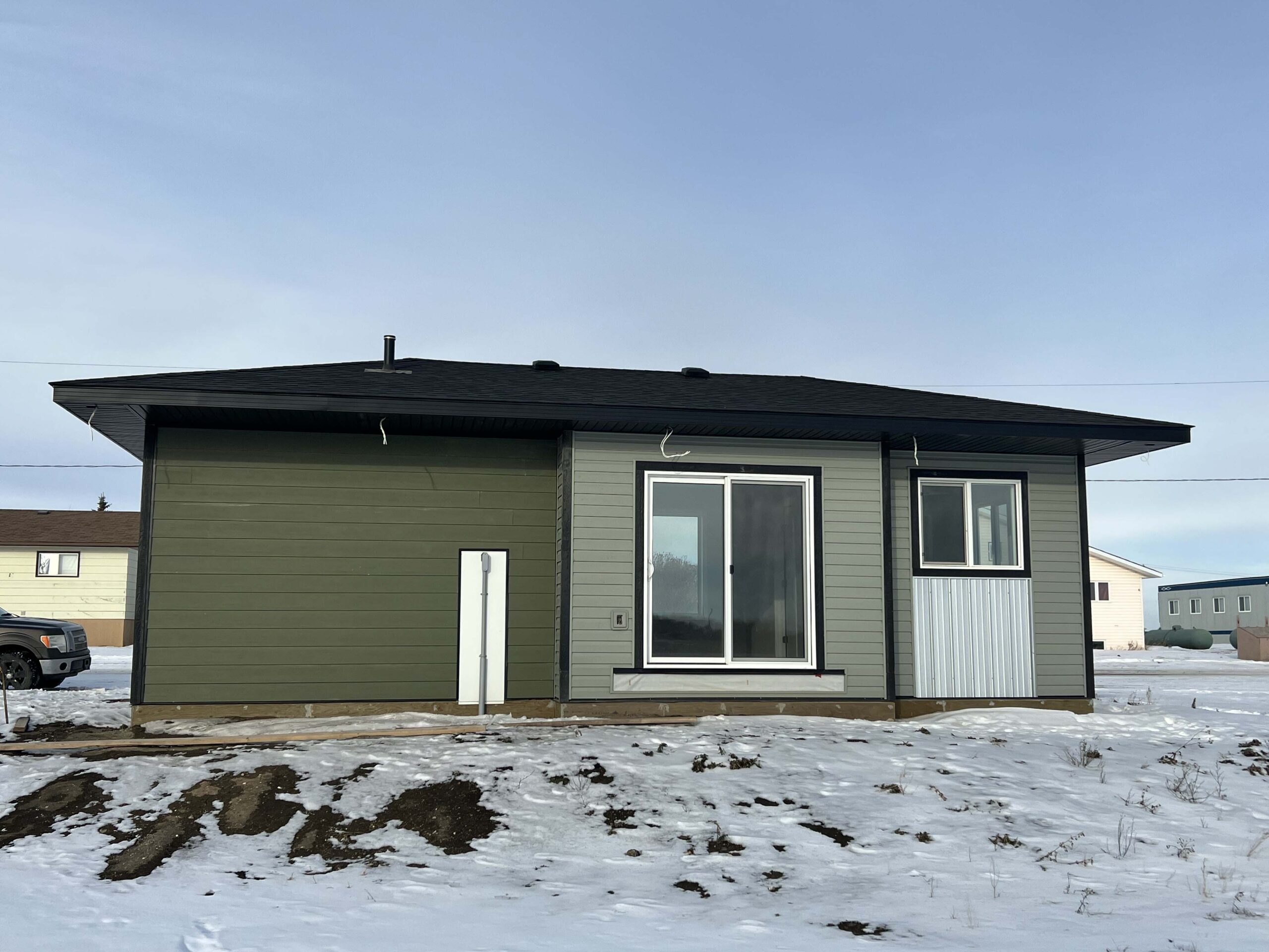 A modern single-story house with green and light gray vinyl siding, large windows, and a black roof, surrounded by snow.