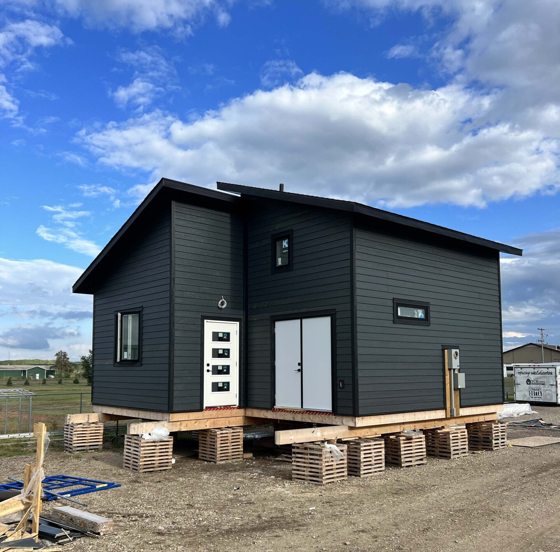 A modern tiny home under construction with dark gray siding, white doors, and elevated on temporary wooden blocks.