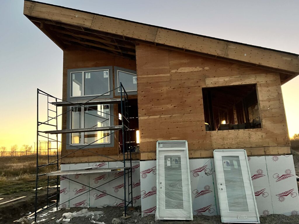 Modern house under construction with exposed plywood framing, large windows installed, and doors ready for installation. Scaffolding is set up for exterior work.