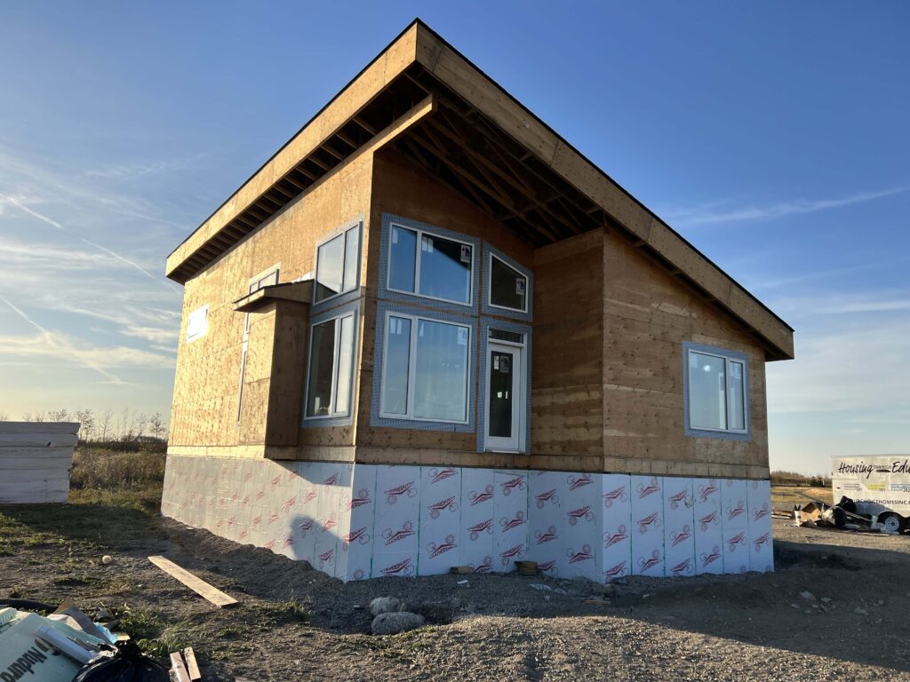 Modern-style home under construction with plywood exterior, newly installed windows, and partial foundation insulation, set against a clear blue sky.