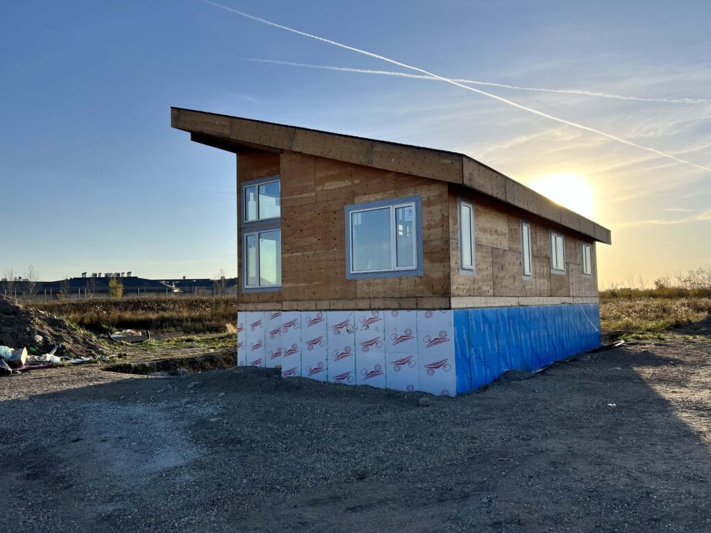 Modern single-story home under construction with plywood siding, large windows, and insulated foundation, set against a sunset backdrop.