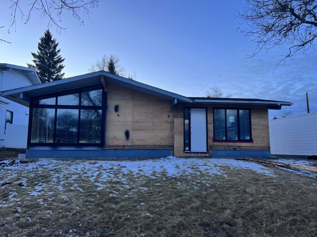 Mid-century modern home under renovation with exposed plywood exterior, large black-framed windows, and a new white door, set against a snowy lawn.