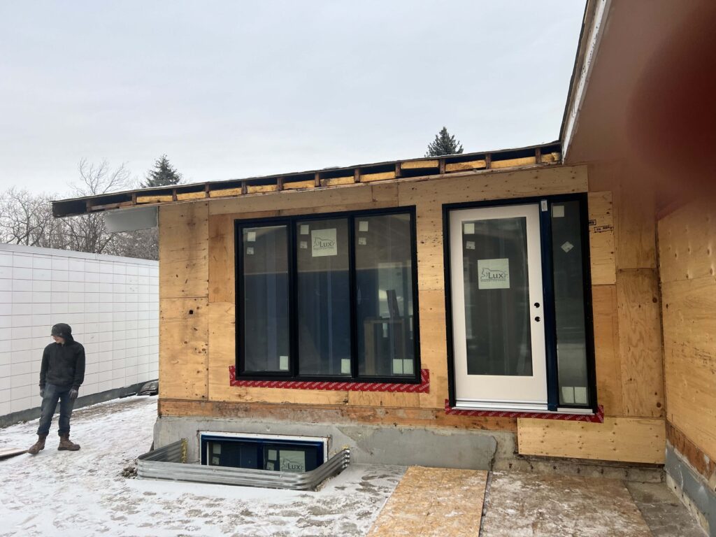 Mid-century modern home under renovation with plywood exterior, newly installed black-framed windows, and a white door, featuring a worker in the foreground on a snow-dusted ground.