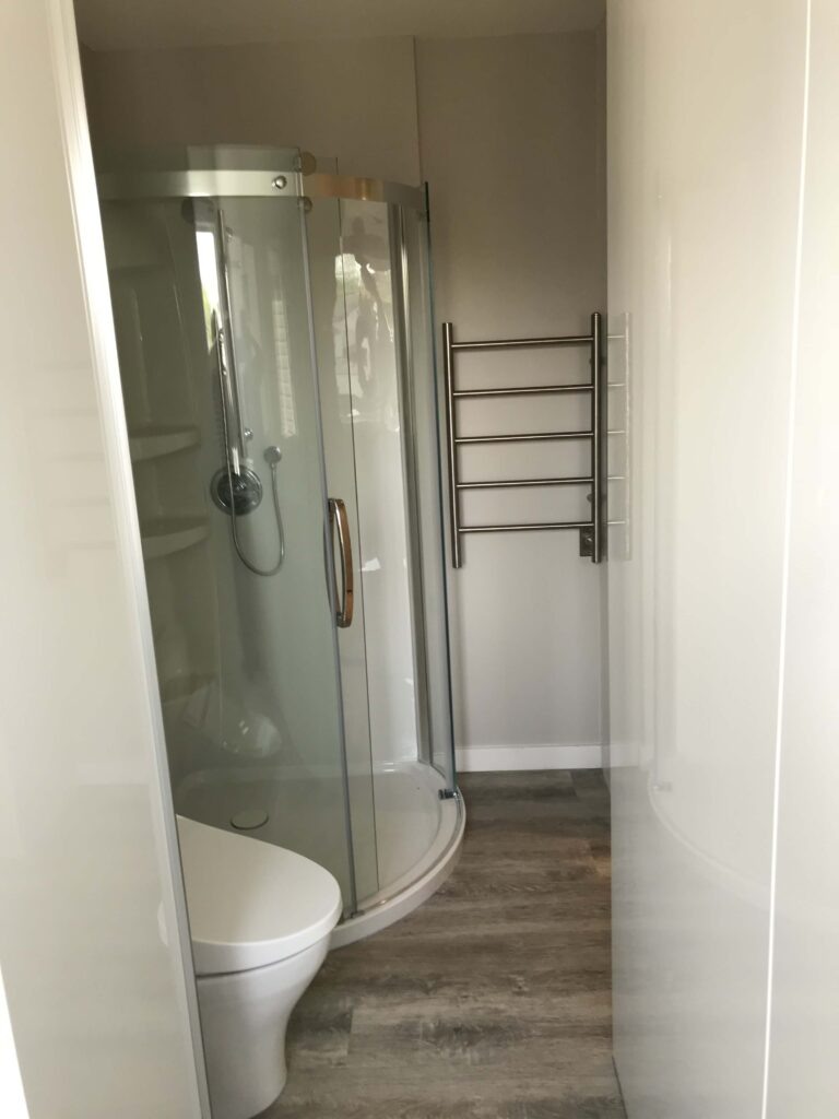 A modern bathroom with a compact curved glass shower, sleek heated towel rack, and wood-style flooring.