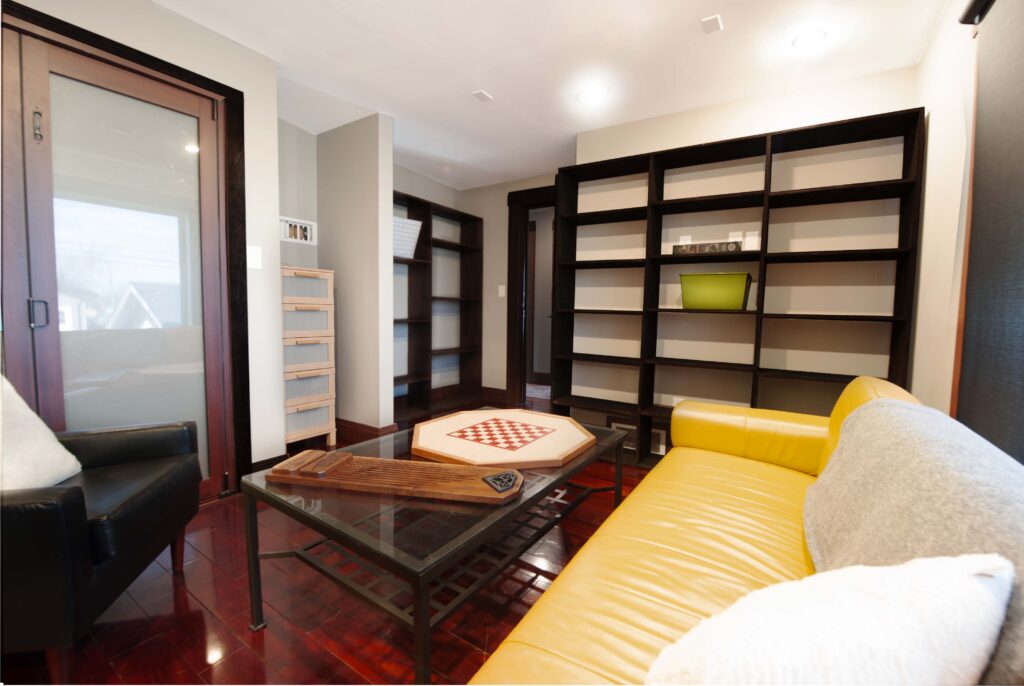 Renovated den with modern built-in shelving, yellow leather sofa, and frosted glass door by Alberta Builder Services.