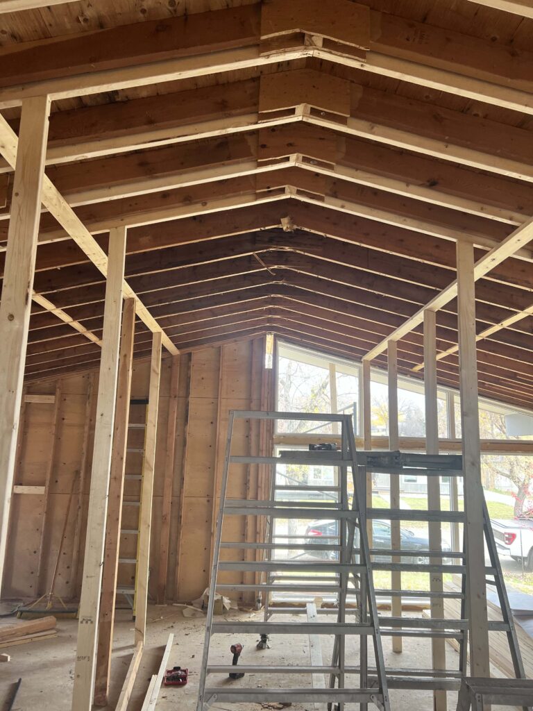 Interior of a renovation project with exposed wooden trusses, open framing, and ladders against a backdrop of windows.