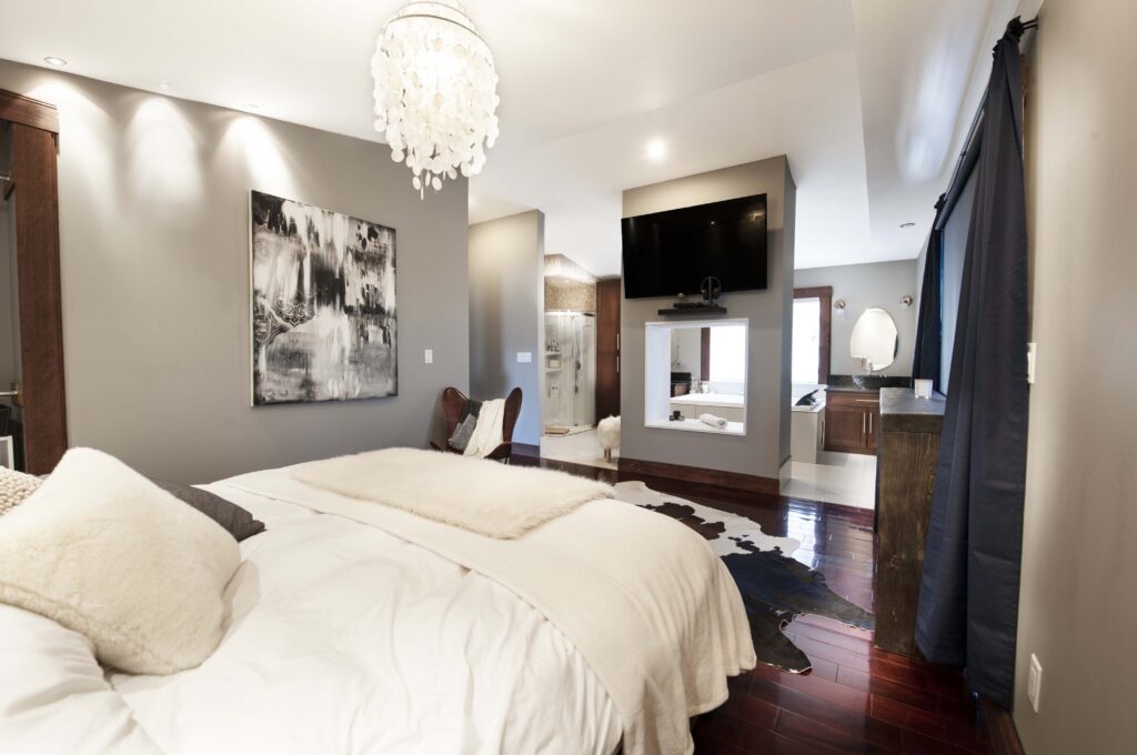 Modern master bedroom with chandelier, open ensuite, TV wall mount, and cowhide rug.