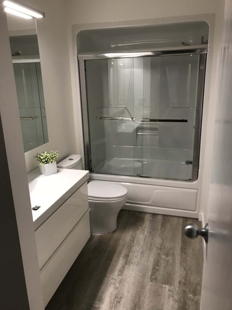 Modern bathroom renovation featuring a sleek vanity, compact toilet, wood-style flooring, and a glass-enclosed bathtub.