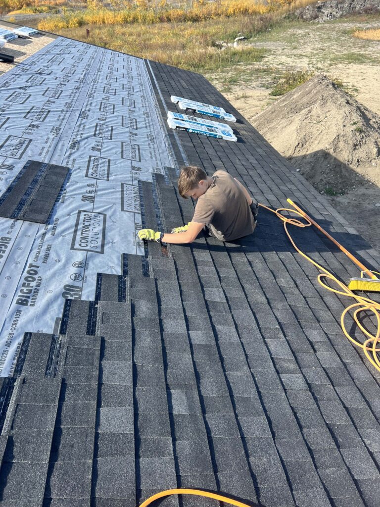 A worker installing asphalt shingles on a residential roof, with underlayment visible in the background.