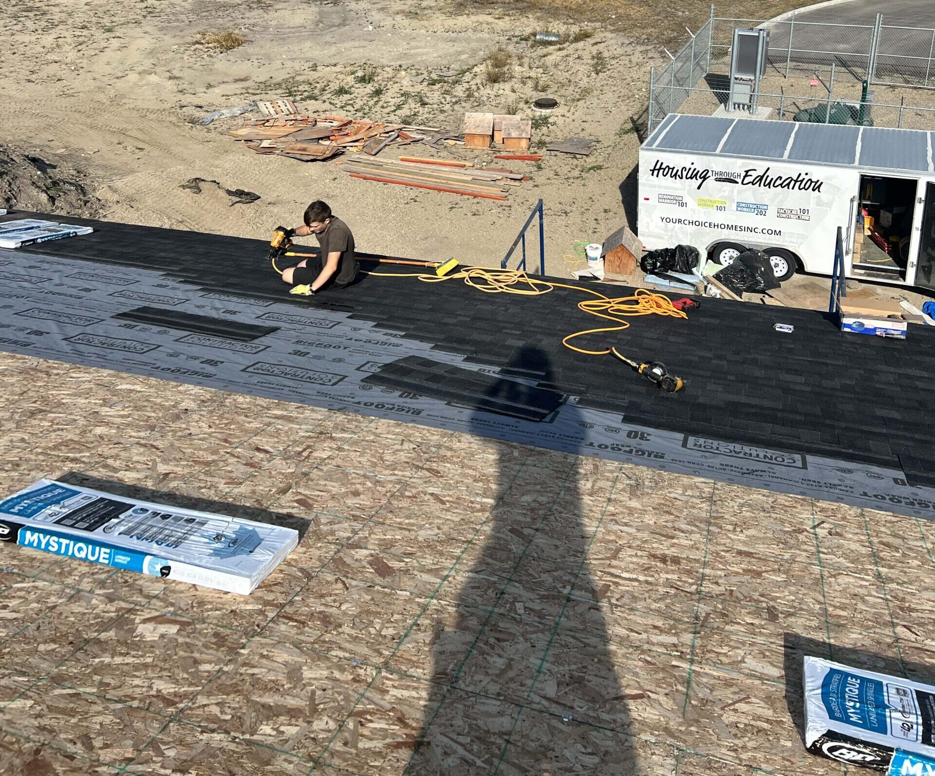 Roofing installation in progress, with a worker laying shingles on a roof under a clear blue sky.