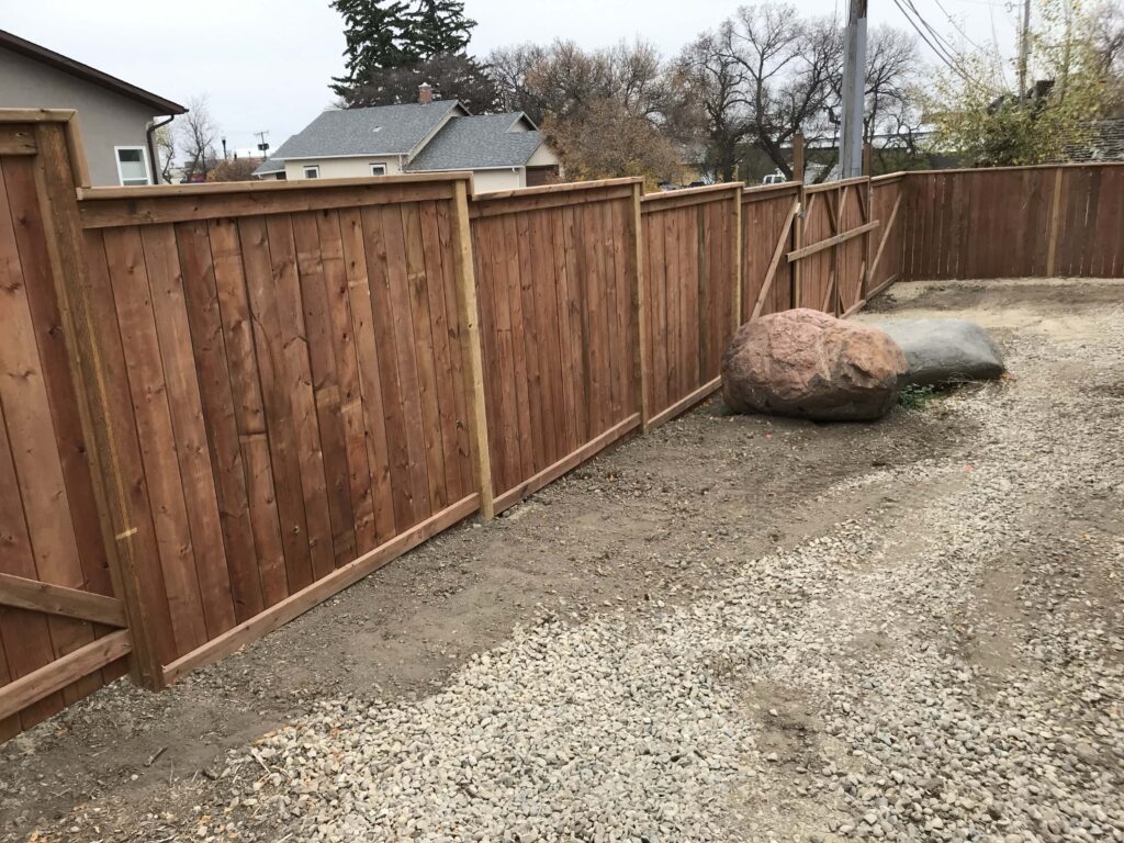 A custom wooden fence with a natural finish, designed and installed by Alberta Builder Services, featuring a clean and durable design along a gravel driveway with decorative boulders.