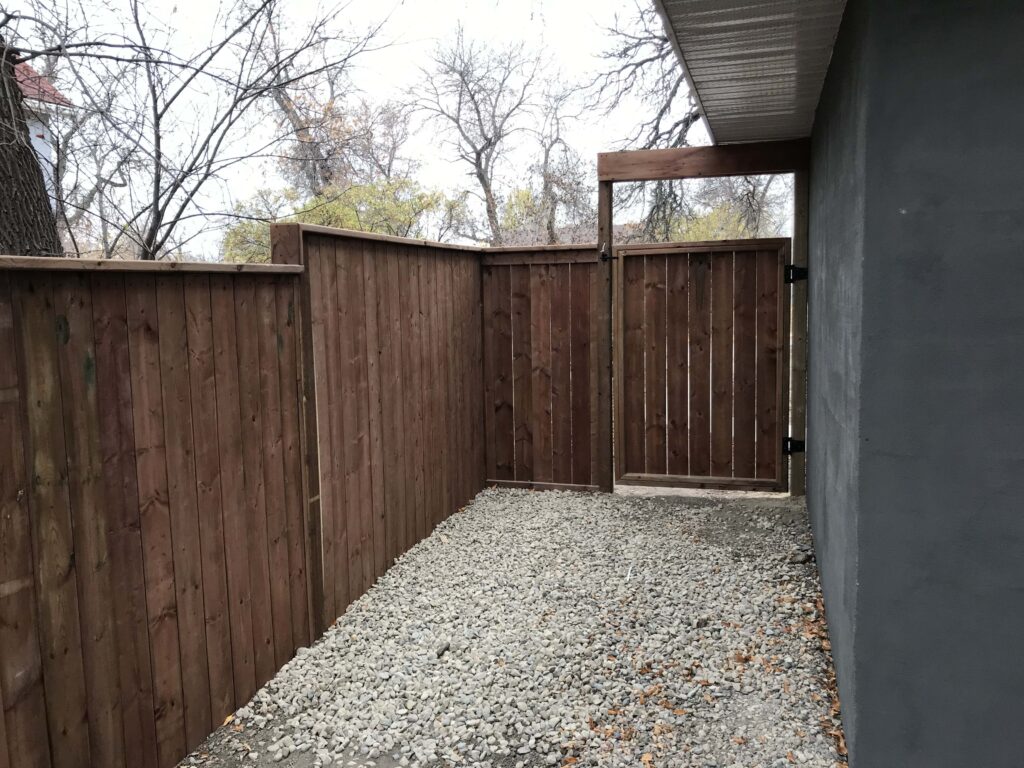 A custom wooden side gate with vertical panels, built by Alberta Builder Services, featuring a sturdy design along a gravel pathway for added privacy and accessibility.