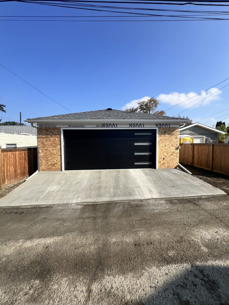 Completed detached garage in Edmonton with modern black overhead door, finished concrete apron, and shingled roof by Alberta Builder Services Ltd.