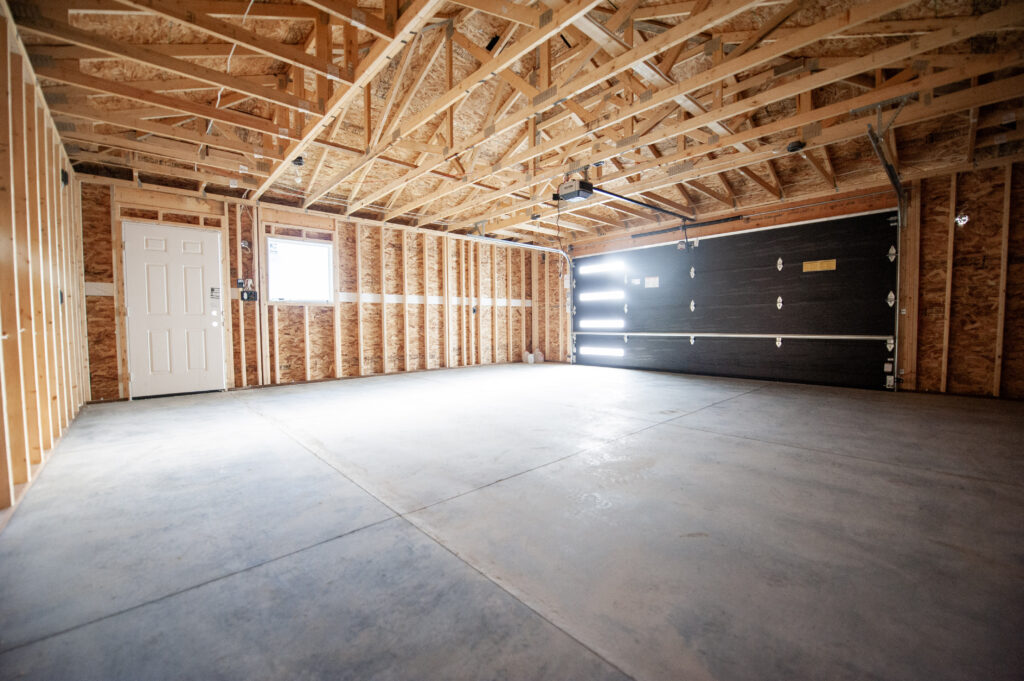 Interior of a newly built unfinished garage with exposed framing, truss roof system, concrete slab floor, and an installed overhead garage door.