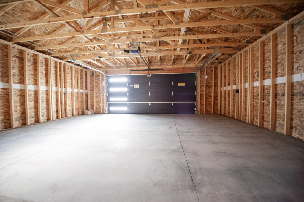 Interior of a newly constructed unfinished double garage with exposed framing, open truss ceiling, concrete slab floor, and an insulated overhead garage door.