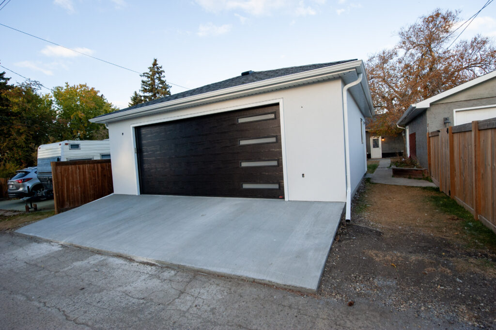 Modern finished detached garage with Sandstorm stucco exterior, a dark overhead door with glass inserts, and a newly poured concrete driveway.