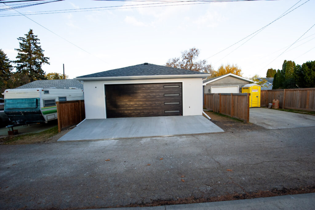 Modern detached garage with an insulated overhead door, fresh concrete driveway, and acrylic stucco exterior in a residential alley.