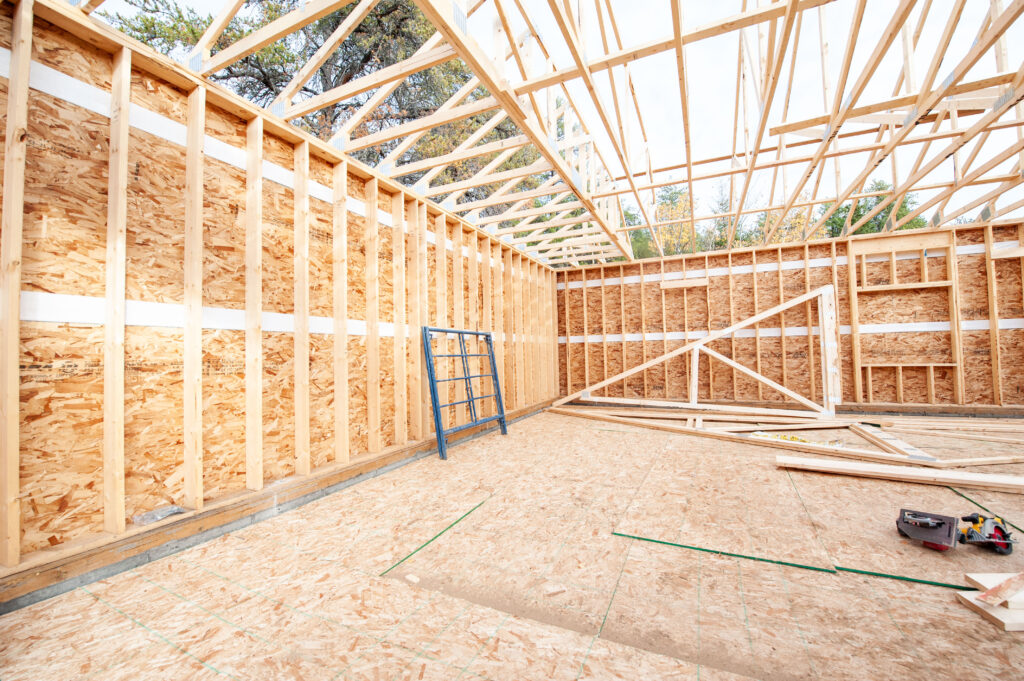 Interior view of an attached garage under construction with framed walls, OSB sheathing, roof trusses installed, and tools on the subfloor.