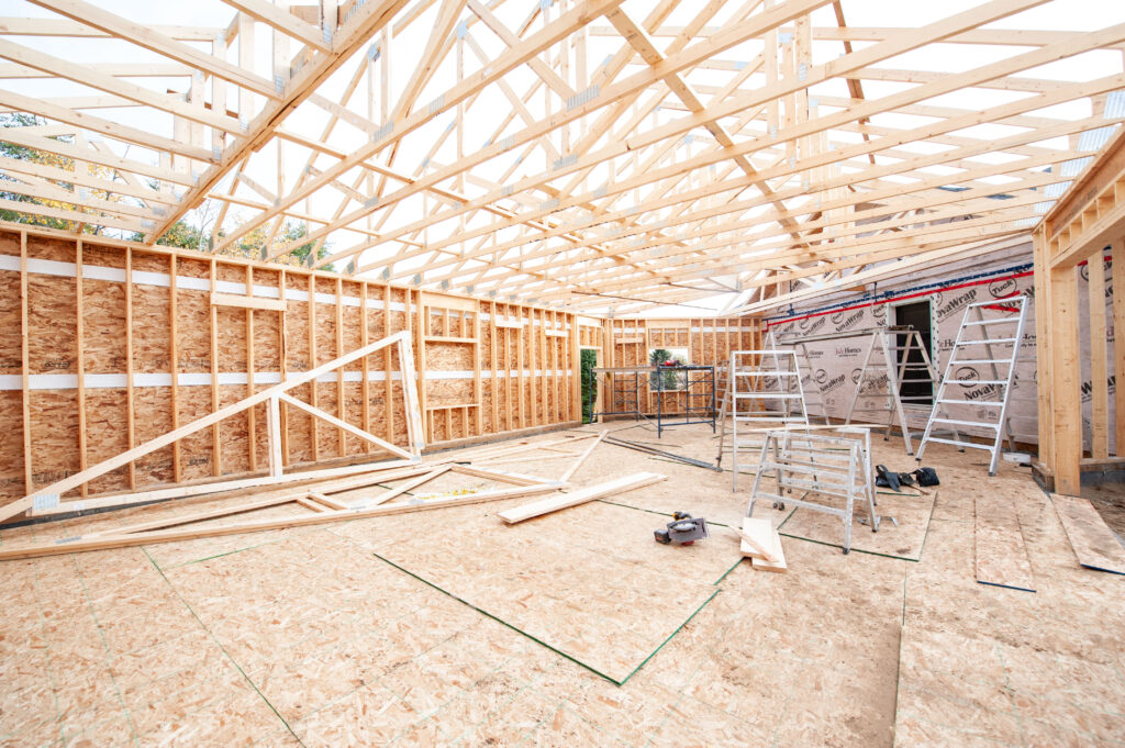 Interior construction view of an attached garage with roof trusses installed, framed walls, OSB sheathing, ladders, scaffolding, and tools on the subfloor.