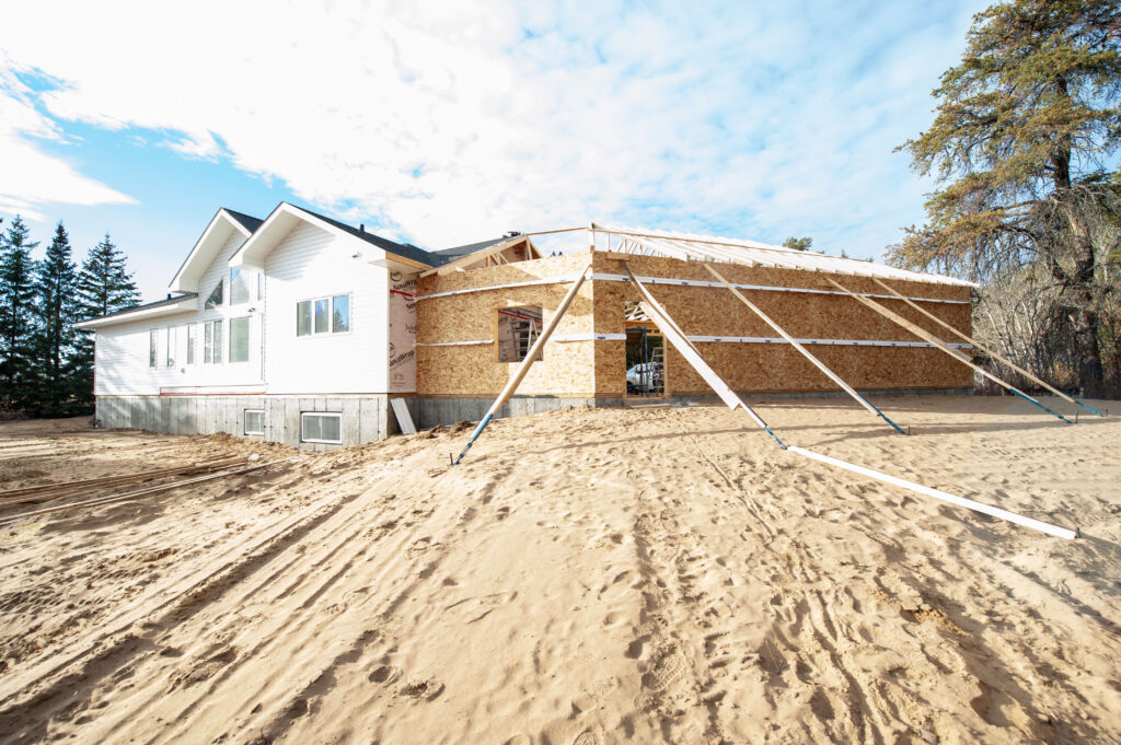 Exterior view of an attached garage under construction with framed walls, OSB sheathing, roof trusses installed, and temporary bracing beside a finished home on a sandy lot.