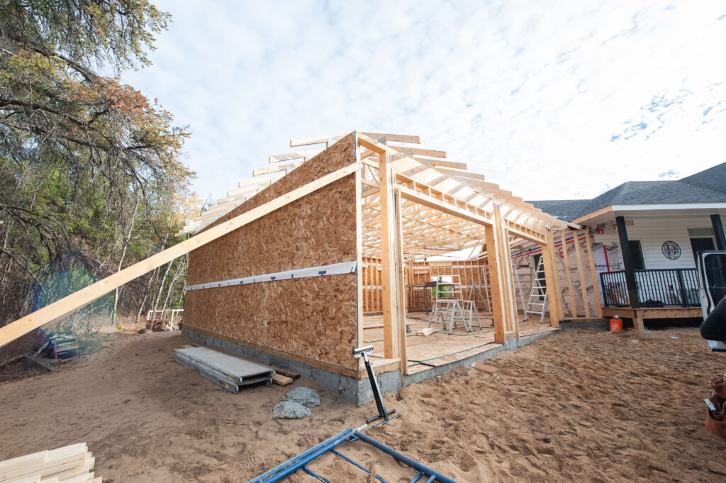 Exterior view of attached garage framing with OSB sheathing, roof trusses installed, and temporary bracing beside an existing home during construction.