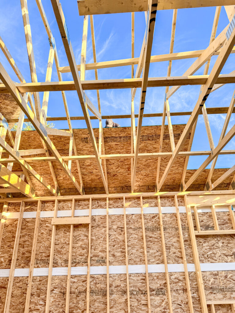Worker installing roof sheathing on an attached garage under construction, viewed from inside the framed structure with exposed trusses.