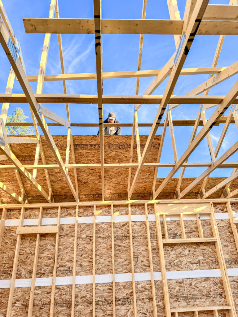 Worker installing roof sheathing on an attached garage under construction, viewed from inside the framed structure with exposed trusses.