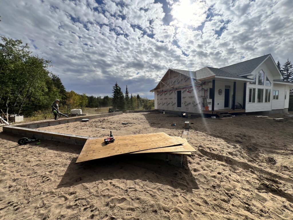 A construction site with a partially built garage foundation beside a finished home, surrounded by sand and trees, under a cloudy sky.
