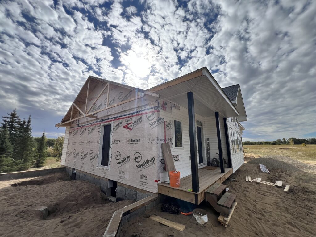 Partially finished home exterior with house wrap and porch under construction on a sandy lot with a cloudy sky overhead.
