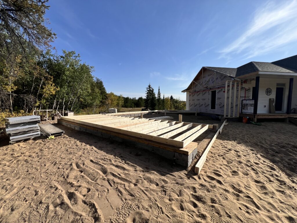 Pre-assembled framed walls laid out on the foundation beside a new home under construction on a sandy lot under a clear sky.