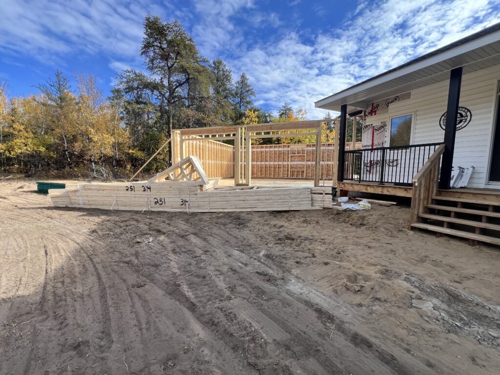 Lumber and truss materials staged on a residential construction site beside a partially framed attached garage under a bright blue sky.
