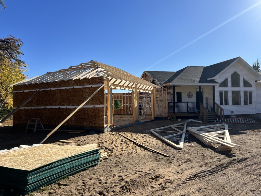Partially framed attached garage with installed roof trusses beside a finished home, with construction materials staged on a sandy worksite.