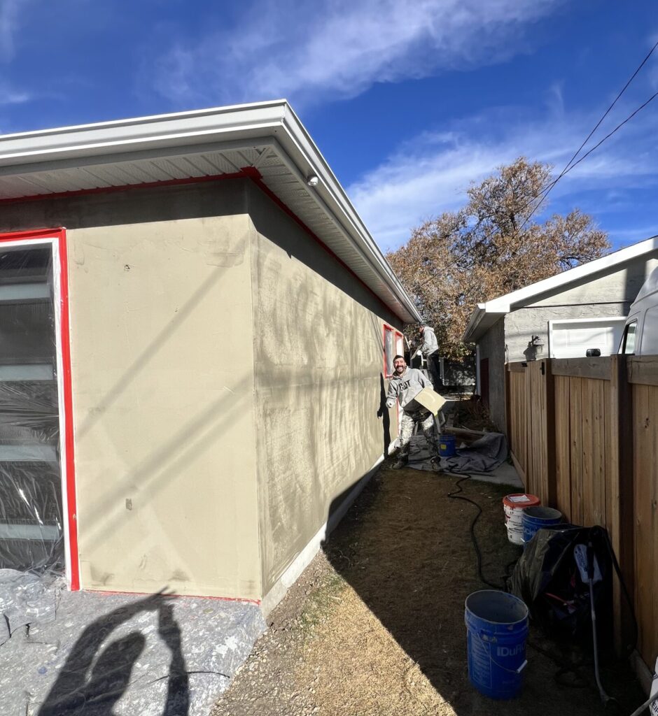 Installer applying an acrylic stucco base coat to the side of a garage, with tools and buckets along the fence line.
