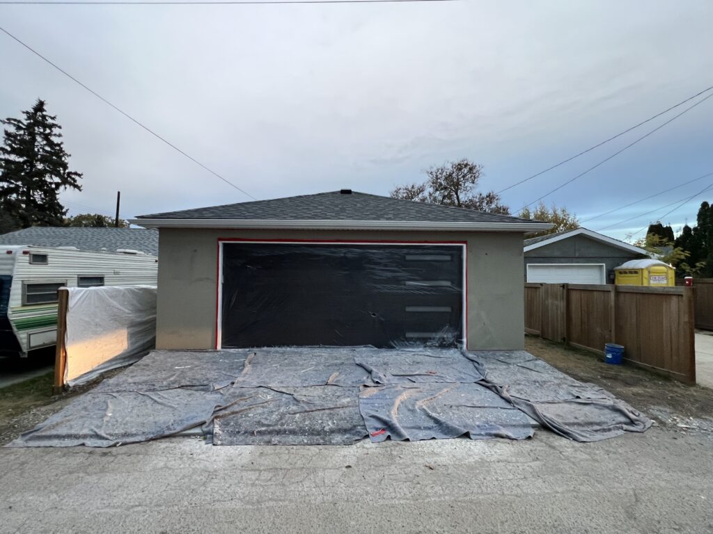 Side view of a detached garage with a freshly applied acrylic stucco base coat and taped openings during exterior finishing.