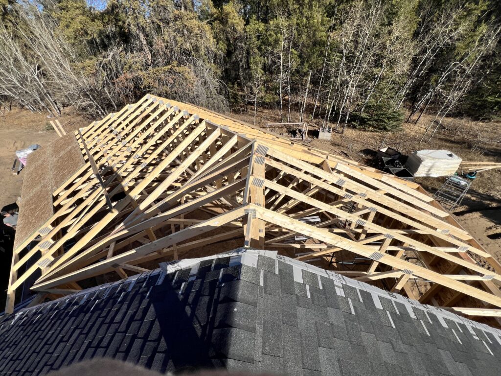 Aerial view of roof trusses installed on an attached garage under construction beside an existing home, with partial roof sheathing and shingles visible.