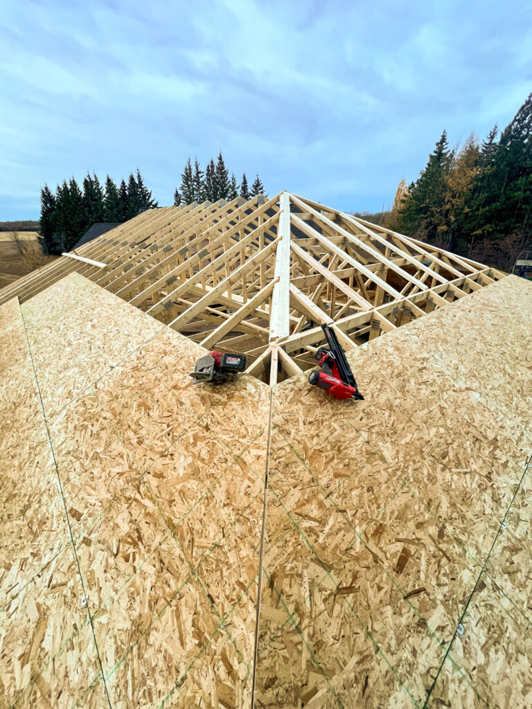 View from the roof of an attached garage under construction with OSB roof sheathing installed, exposed trusses below, and construction tools resting on the roof.