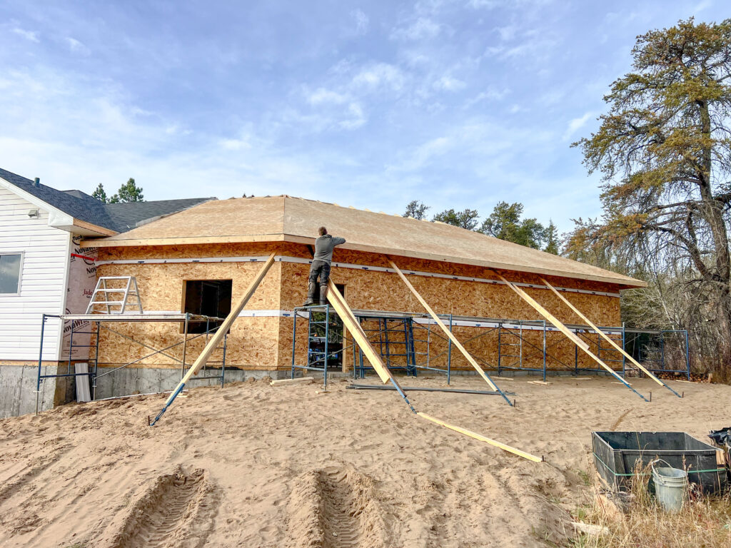 Exterior view of an attached garage under construction with roof sheathing installed, temporary bracing in place, scaffolding, ladders, and a worker completing roofing preparation.