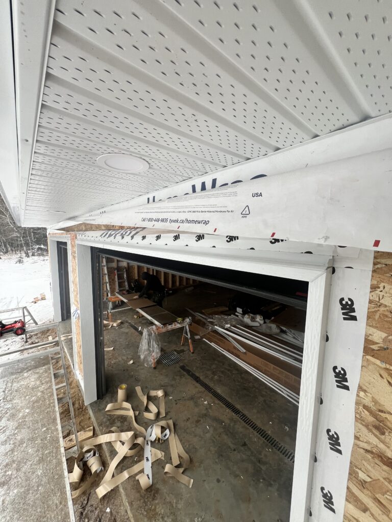 Close-up view of garage exterior during construction, showing installed soffit, garage door trim flashing, house wrap, and unfinished interior workspace.