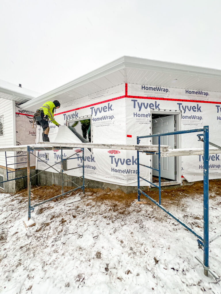 Worker installing exterior house wrap on a garage under construction in snowy conditions, with scaffolding and Tyvek wrap visible.