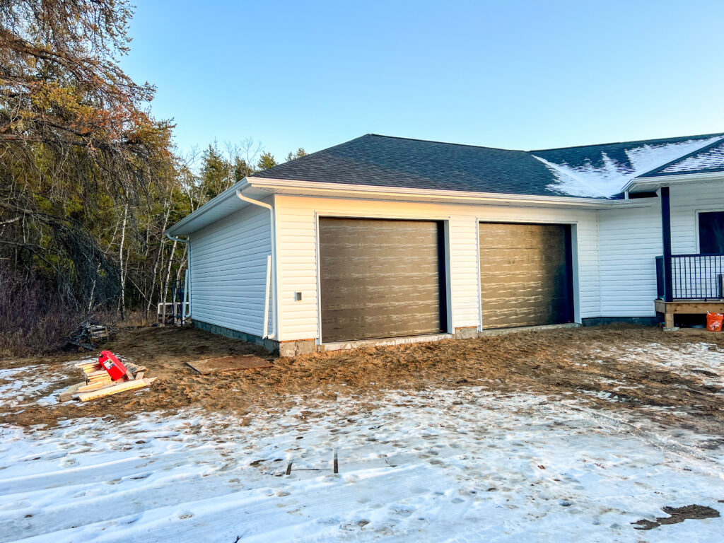 Completed two-door attached garage build with new white vinyl siding and installed garage doors beside an existing home in a snowy yard.