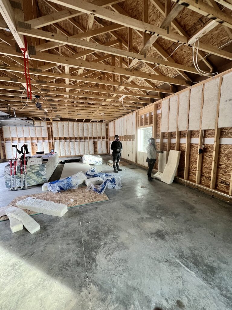 Two workers installing insulation inside a framed garage with exposed trusses and construction materials on the floor.