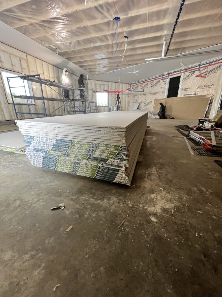 Workers installing drywall in an insulated garage with stacked Sheetrock and scaffolding.