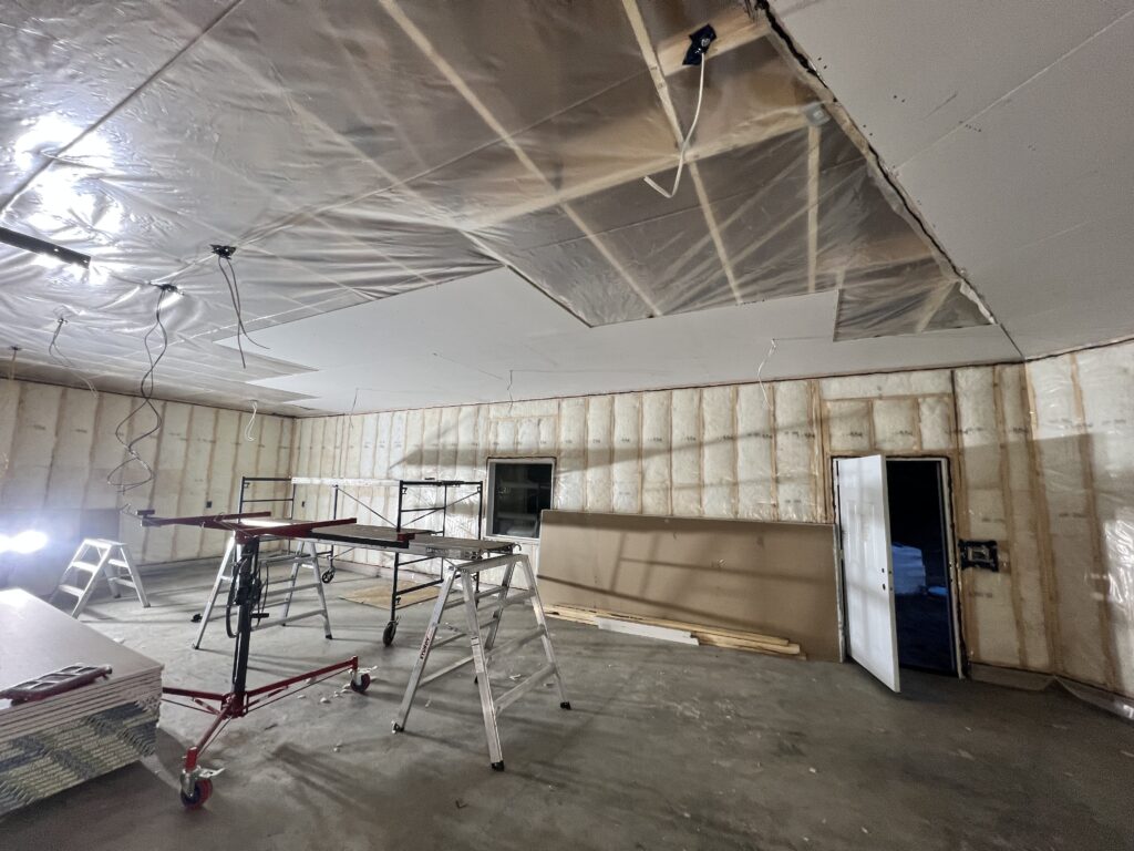 Drywall installation in progress inside an insulated garage with ladders, scaffolding, and a drywall lift.
