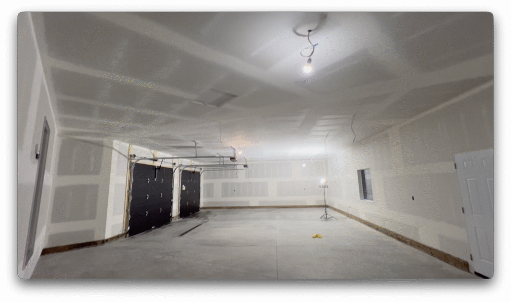 Finished garage interior with drywall completed, concrete floor, black overhead doors, and temporary lighting.