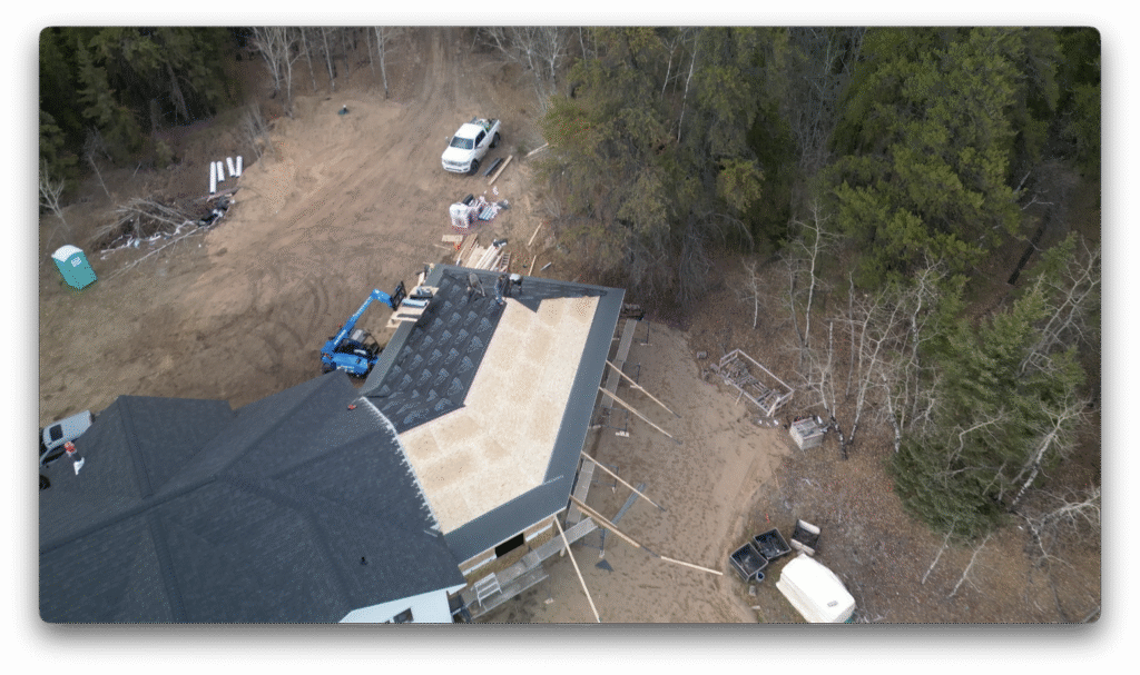A drone photo showing workers installing roofing underlayment on a new home build, with equipment, scaffolding, and construction materials surrounding the site.