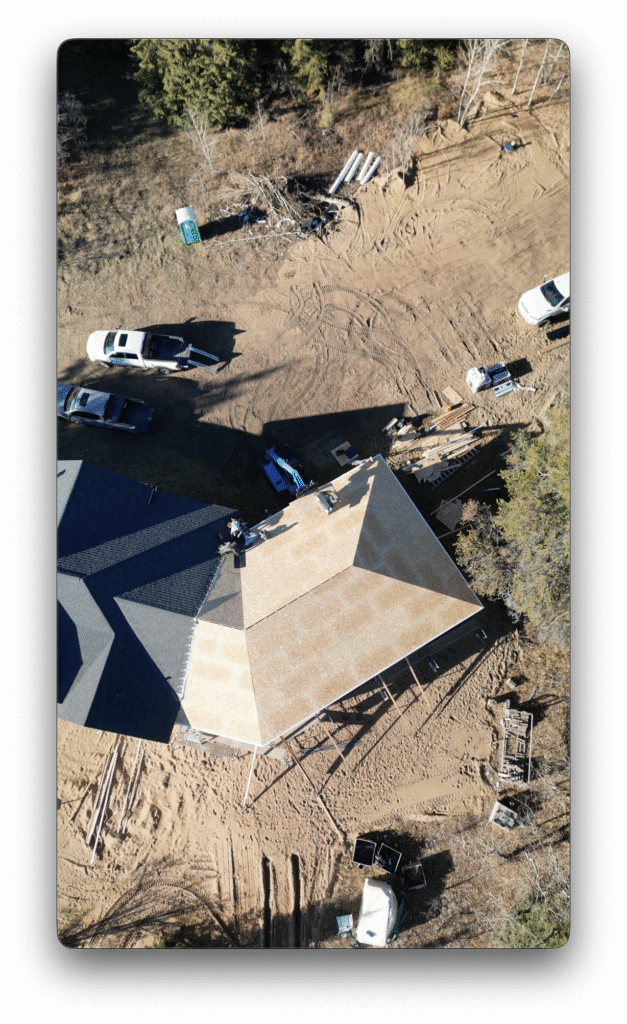 A drone photo showing roof construction on a house, with workers installing roofing materials and vehicles, equipment, and building supplies spread across the surrounding dirt site.