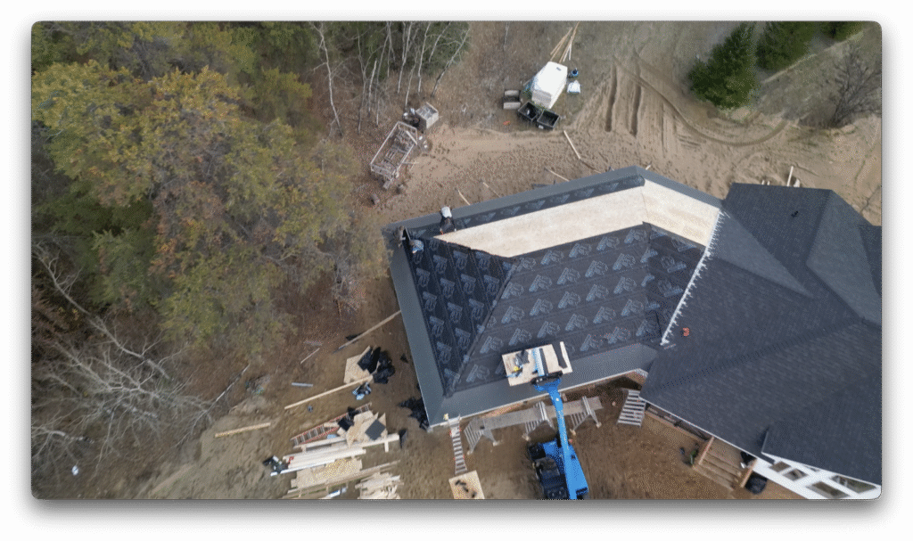 A drone image showing workers installing roofing underlayment on a partially completed roof, with construction equipment and materials scattered around the work site.
