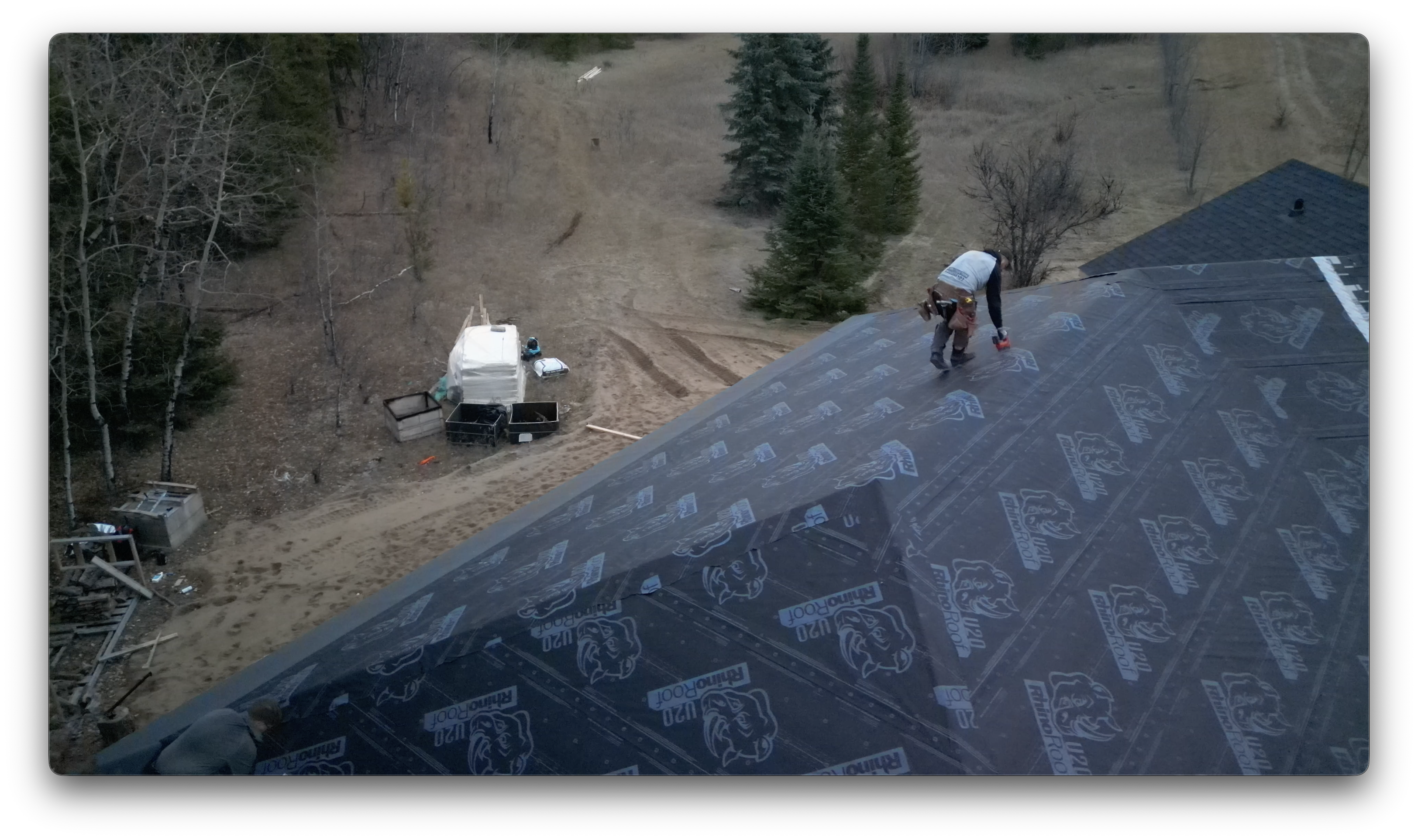 A worker installs roofing underlayment on a steep roof while construction materials and wooded surroundings are visible below.